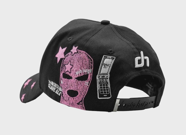 Op de afbeelding: Zwarte baseballcap met roze borduursels. Het heeft een roze bivakmuts-afbeelding met sterren, een klaptelefoon en de tekst "CLAVE ALTO". De achterkant heeft een wit "dh"-logo en een verstelbare band.