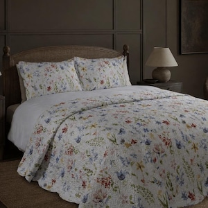 May include: A white quilt and two matching pillow shams featuring a colourful botanical print of small flowers in shades of blue, red, yellow, and green. The bedding set has a textured, quilted surface with a scalloped edge along the bottom hem.