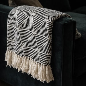 May include: A monochrome throw blanket with a geometric pattern and cream-coloured fringe is draped over a dark green sofa. The blanket's design features repeating triangles.