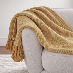 May include: A mustard yellow throw blanket featuring a woven herringbone pattern and long, thick tassels along the edge. The fabric has a textured appearance with white thread accents integrated into the weave, draped over a light-coloured armchair.