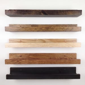 May include: Five horizontal wooden wall shelves arranged in a vertical stack. Each shelf features a rectangular ledge and a back panel. The wood finishes vary in color from dark espresso and medium brown to a light natural tone and black.
