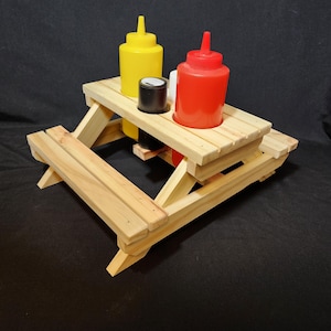 May include: A miniature wooden picnic table with a yellow mustard bottle, a red ketchup bottle, and a small black container. The table is made of light-colored wood and has a classic picnic table design.