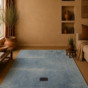May include: A rectangular area rug featuring a faded light blue surface with a visible central seam. The rug includes a small, dark rectangular patch in the center and two smaller, lighter blue circular patches positioned on the right and bottom sides.