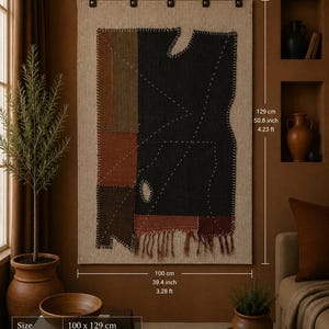 Puede incluir: Un tapiz rectangular que presenta un gran panel de tela negra con contornos geométricos cosidos en blanco, acentuado por parches de color marrón y óxido en el lado izquierdo. Mide 100 cm por 129 cm y cuelga de una varilla de madera.