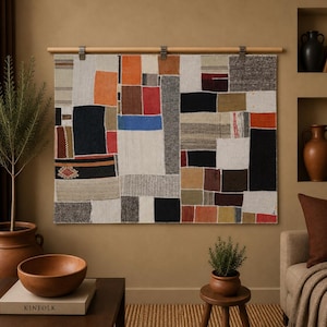 Peut inclure: Une tenture murale rectangulaire présentant un motif en patchwork composé de diverses textures et couleurs de tissus, notamment orange, rouge, bleu, noir, blanc et gris. Le textile est suspendu à une tringle en bois clair avec trois clips.