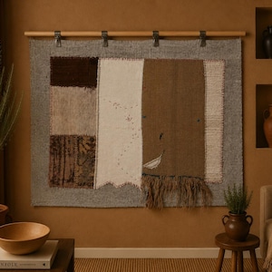 May include: A rectangular wall hanging featuring a patchwork of textured fabric panels in shades of brown, cream, and gray. The textile is suspended from a light wood rod with metal clips and includes visible stitching and a fringed bottom edge.