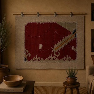 Puede incluir: Un tapiz rectangular que presenta un gran panel de tela de color rojo intenso con patrones geométricos y bordes cosidos, montado en una barra de madera clara con cuatro clips metálicos, sobre una pared de textura neutra.