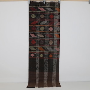May include: A long, rectangular textile runner featuring a dark brown and black base with a grid of vertical and horizontal stripes. The fabric is decorated with small, repeating diamond-shaped patterns in red, orange, yellow, and white thread.