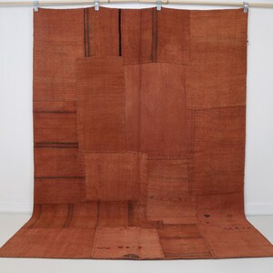 May include: A large rectangular rug composed of multiple stitched panels in varying shades of terracotta and burnt orange. The textile features a visible patchwork construction with dark brown linear accents and subtle geometric patterns.
