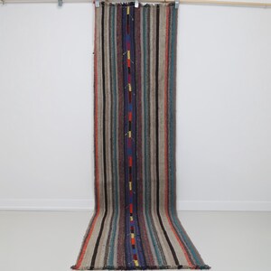 May include: A long, narrow runner rug featuring a vertical striped pattern in shades of beige, brown, gray, and blue. A central dark blue stripe is accented with small, repeating rectangular blocks of bright yellow, red, and orange colors.
