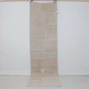 May include: A rectangular, beige patchwork rug with a neutral color palette. The rug is made of several rectangular pieces sewn together. The rug is hanging from a wooden rod against a white wall.