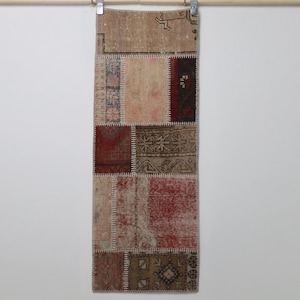 May include: A rectangular, patchwork rug with a variety of colors including beige, red, and brown. The rug is made of several rectangular pieces sewn together. The rug is hanging from a wooden rod with metal clips.
