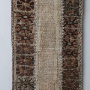 May include: Rectangular rug with a central beige panel flanked by patterned brown panels. The rug is made of woven material and has a stitched border. It is suspended by metal clips from a wooden rod.