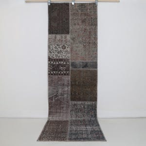 May include: A rectangular, patchwork rug with a variety of muted colors, including brown, gray, and beige. The rug is made up of several rectangular patches with different patterns and textures. The rug is hanging from a wooden rod.