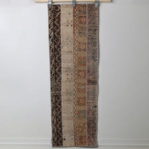 May include: A rectangular, vintage-style runner rug with a patchwork design. The rug features vertical stripes in various colors, including brown, beige, and muted orange, with decorative patterns. It is hanging against a white wall.
