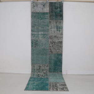 May include: A long, rectangular rug with a patchwork design. The rug features various shades of teal, gray, and faded patterns. It is hanging from a metal rod against a white wall. The rug's design is a mix of geometric and abstract shapes.