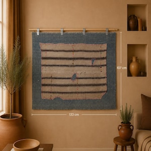 Puede incluir: Un tapiz rectangular con un panel central de tela beige con rayas horizontales marrones y dos pequeños agujeros, bordeado por un marco de tela azul oscuro. Está suspendido de una varilla de madera con cuatro clips metálicos, midiendo 133 cm por 107 cm.