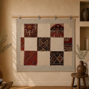 Op de afbeelding: Een rechthoekige wandhanger met een patchworkontwerp van crèmekleurige stoffen vierkanten die afwisselen met vierkanten van donkerrood, bruin en zwart textiel met patronen. Het stuk hangt aan een lichte houten roede met metalen clips.