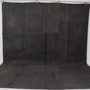 Puede incluir: Una alfombra grande de color gris oscuro con un diseño de retazos. La alfombra es rectangular y parece estar hecha de un material tejido. Cuelga de una barra de madera con pinzas metálicas. La alfombra se muestra sobre un fondo blanco.