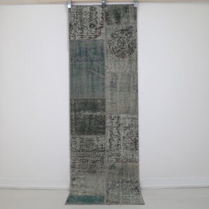 Può includere: Un tappeto rettangolare patchwork dall'aspetto vintage. Il tappeto presenta un mix di colori tenui, tra cui verde acqua, grigio e beige, disposti in motivi geometrici. Il tappeto è appeso a una parete bianca.