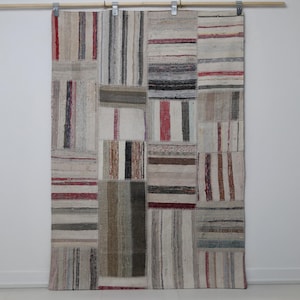 May include: A patchwork rug in neutral tones, with rectangular and striped designs in beige, grey, and red. The rug is suspended from a wooden rod with metal clips, suitable as a decorative wall hanging. The rug's dimensions are in centimetres.