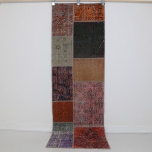 May include: A rectangular patchwork rug with a variety of colors and patterns. The rug is made up of rectangular patches in shades of red, orange, green, brown, and purple. The rug is hanging against a white wall.