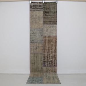 Può includere: Un tappeto rettangolare patchwork con una varietà di colori tenui, tra cui marrone, beige e verde acqua. Il tappeto è composto da diversi pezzi rettangolari cuciti insieme. Il tappeto è appeso contro un muro bianco.