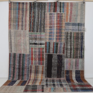 Può includere: Un grande tappeto rettangolare composto da vari pezzi di tessuto cuciti insieme in un design patchwork. Il tappeto presenta una tavolozza di colori variegata che include tonalità di grigio, blu, rosso, marrone e nero, con cuciture visibili.