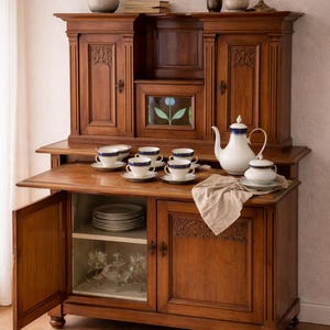Puede incluir: Un mueble antiguo de madera oscura con una alacena, puertas abiertas y una exhibición de tazas de té, platillos y una tetera. El mueble tiene detalles tallados y una sección con frente de vidrio. Un jarrón de trigo y una vela encendida están encima.