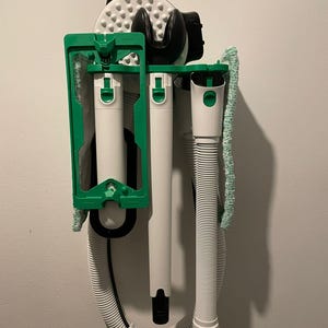 Puede incluir: Un juego de herramientas de limpieza blanco y verde, montado en una pared. El juego incluye una unidad portátil, dos componentes cilíndricos blancos y una manguera flexible. Los componentes se sujetan con un marco verde.