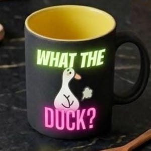 May include: A matte black ceramic mug with a bright yellow interior and a matching black handle. The front features a white cartoon duck illustration with a small grey cloud, accompanied by the text WHAT THE in light green and DUCK? in pink.