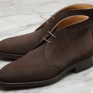 Puede incluir: Un par de botines chukka de ante marrón. Los botines tienen un diseño clásico con punta redondeada, cierre de cordones y tacón bajo. Están hechos de ante suave y texturizado y tienen una suela marrón oscuro.