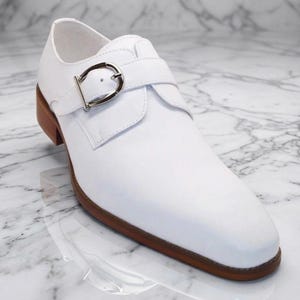 Könnte beinhalten: Ein weißer Lederschuh mit silberner Schnalle. Der Schuh hat eine braune Sohle und einen Absatz. Das Design ist ein klassischer Monk-Strap-Stil, geeignet für formelle oder halbformelle Anlässe.