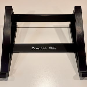 Può includere: Supporto nero con la scritta "Fractal FM3" in bianco. Il supporto ha una forma rettangolare con due gambe angolate e una barra orizzontale. Il supporto è progettato per contenere un dispositivo elettronico.