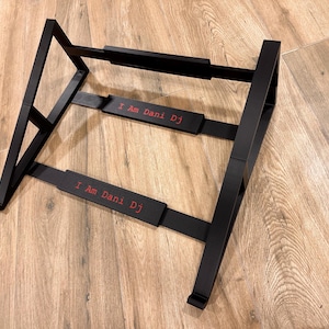 Può includere: Supporto per DJ in metallo nero con la scritta "I Am Dani Dj" in rosso. Il supporto ha un design angolato con due supporti orizzontali ed è progettato per contenere apparecchiature per DJ. Il supporto è su una superficie di legno.