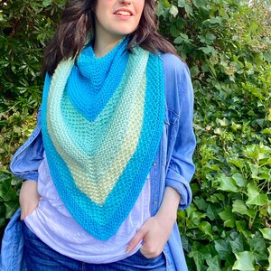 May include: A woman wearing a blue denim jacket and a white t-shirt is standing in front of a green leafy background. She is wearing a blue, green, and white ombre triangular scarf.