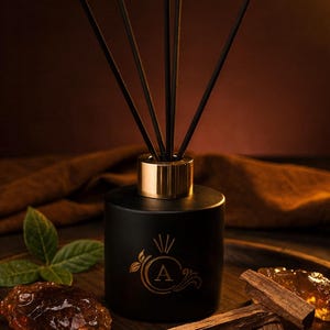 Könnte beinhalten: Ein schwarzer Diffusor für ätherische Öle mit einem goldfarbenen Kragen und schwarzen Stäbchen. Der Diffusor hat ein goldenes Emblem mit dem Buchstaben "A" und ist von Blättern umgeben. Er steht auf einem Holztablett mit Holzstücken und bernsteinfarbenen Kristallen.