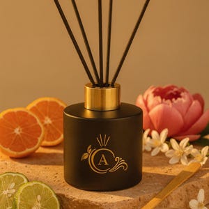 Könnte beinhalten: Ein schwarzer Diffusor für ätherische Öle mit goldfarbenem Kragen und Stäbchen. Der Diffusor hat ein goldfarbenes Monogramm und ist von geschnittenen Orangen, Limetten und einer rosa Blume umgeben. Ein kleiner Holzspatel und eine Schale mit Pulver sind ebenfalls vorhanden.