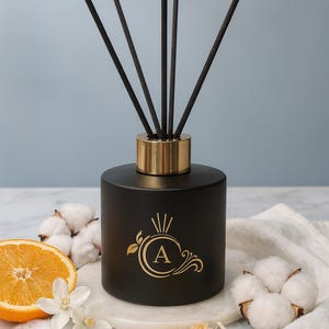 Könnte beinhalten: Schwarzer Diffusor mit goldenen Akzenten und Stäbchen. Die Flasche hat ein goldenes Emblem mit dem Buchstaben "A" und dekorativen Elementen. Eine Orangenscheibe, Baumwolle und weiße Blüten sind um den Diffusor angeordnet.