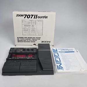 Peut inclure: Un processeur multi-effets Zoom 707II Guitar noir avec une boîte et un manuel d'utilisation assortis. Le processeur comprend plusieurs pédales, un écran d'affichage et divers boutons de commande. La boîte affiche le nom et les caractéristiques du produit.