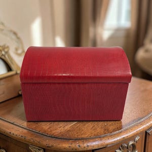 May include: A red, rectangular box with a curved top. The box has a textured, crocodile-like pattern. The box is sitting on a wooden surface with ornate carvings. The box is likely used for storage or decoration.