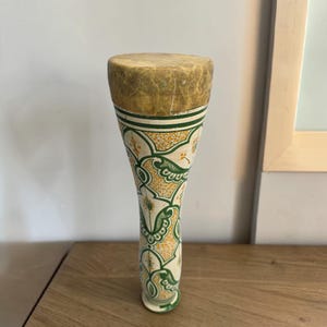 Peut inclure: Un tambour à main haut et élancé avec un corps cylindrique qui s'affine vers la base. La peau du tambour est faite d'une peau animale texturée de couleur beige, tandis que le corps est orné de motifs floraux complexes verts et jaunes.