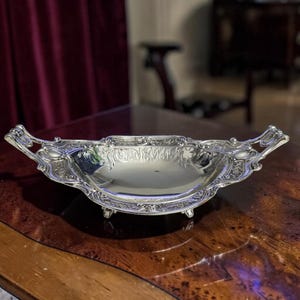 May include: A silver-toned metal serving dish featuring an elongated, shallow bowl with ornate, embossed floral and scrollwork detailing along the rim and handles. The piece is supported by small, decorative feet and has a polished, reflective surface.