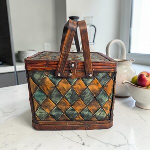 May include: A square picnic basket with a woven pattern in shades of green, brown, and orange. The basket has a wooden handle and a hinged lid. The basket is sitting on a white countertop.