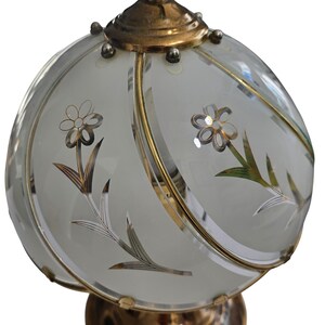 May include: A vintage table lamp with a frosted glass shade featuring floral designs. The shade is adorned with gold-colored trim and accents. The base and top of the lamp are also gold-colored, creating a cohesive and elegant aesthetic.
