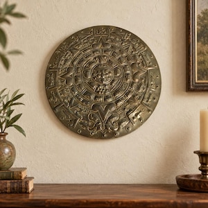 Peut inclure: Une plaque murale circulaire de couleur bronze présentant un relief complexe du calendrier de la pierre du soleil aztèque. La surface métallique affiche un visage central entouré d'anneaux concentriques de glyphes, symboles et motifs géométriques.