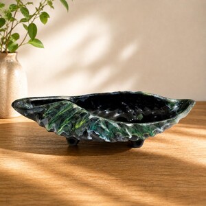 May include: A decorative, leaf-shaped ceramic bowl with a glossy black interior and a textured exterior. The exterior features shades of green, blue, and white. The bowl rests on three small black feet and is suitable for home decor.