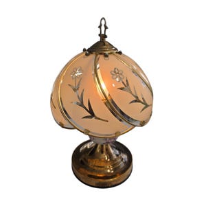 May include: A vintage table lamp with a floral design. The lamp features a peach-colored glass shade with silver floral accents and a brass base. The shade is shaped like a flower, and the lamp is lit.