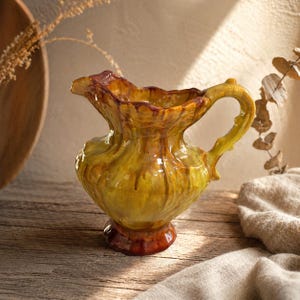Peut inclure: Pichet en céramique fait à la main, avec un glaçage jaune et marron. Le pichet a un bord ondulé unique et une anse incurvée. La base est marron foncé, et le design rappelle la poterie vintage. Le pichet est posé sur une surface en bois.