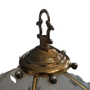 May include: Close-up of a vintage brass and glass pendant light fixture. The fixture features a decorative metal loop for hanging, a brass-colored canopy, and a frosted glass shade. The design includes ornate details and a classic aesthetic.